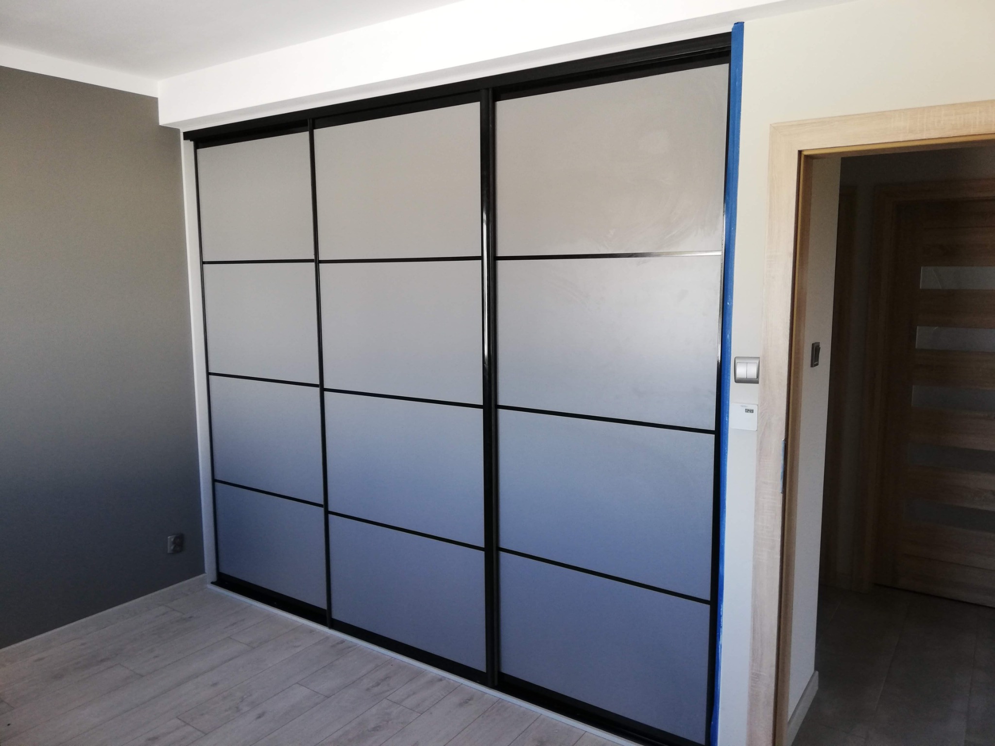 Trzydrzwiowa szafa wnękowa z przesuwnymi, matowymi drzwiami w kolorze szaro-niebieskim, podzielonymi czarnymi listwami na dziewięć prostokątnych pól, widoczna w rogu pokoju na tle szarej ściany...