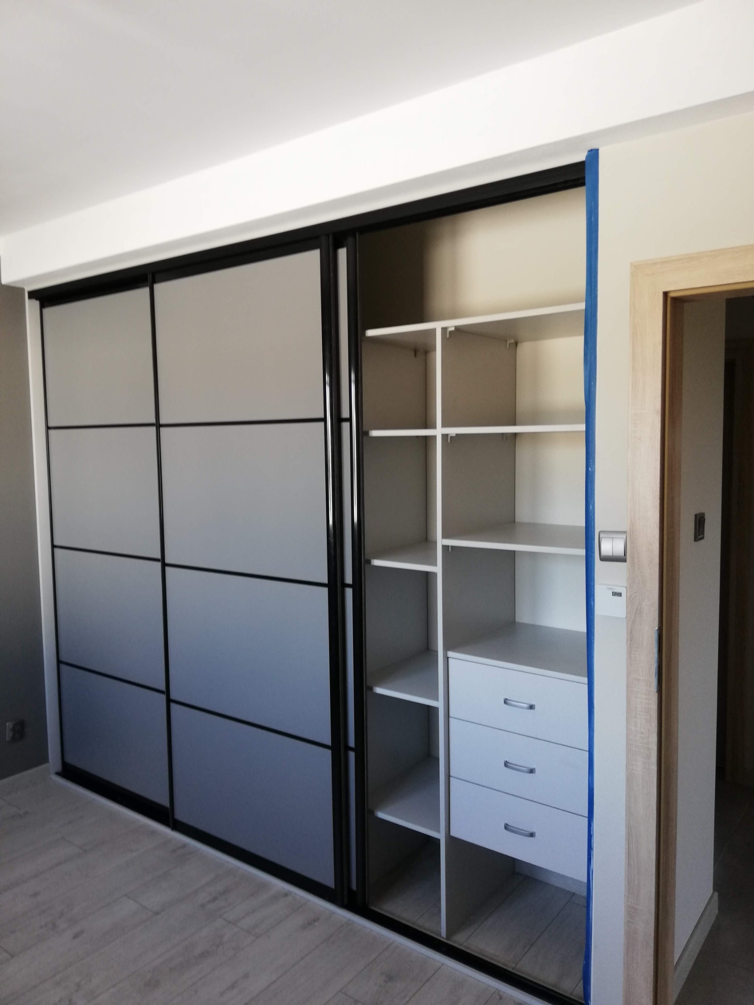 Wnętrze szafy wnękowej z przesuwnymi drzwiami w kolorze szarości, otwarte półki i szuflady, widoczna taśma malarska zabezpieczająca ścianę.