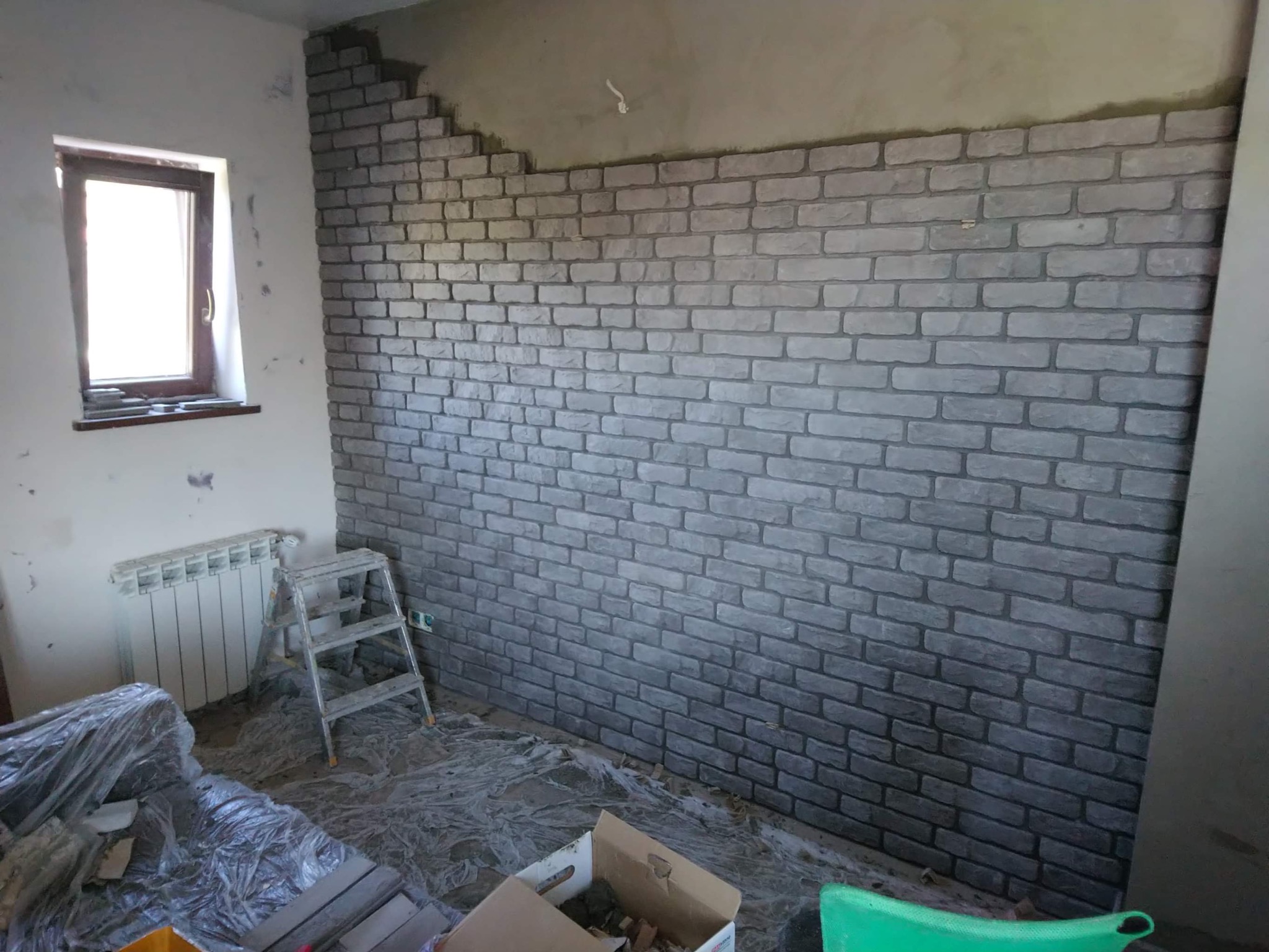 Ściana w trakcie wykańczania z szarych, dekoracyjnych paneli imitujących cegłę, widoczny fragment nieotynkowanej ściany u góry, drabina malarska, zakryte folią meble i kartony na podłodze.