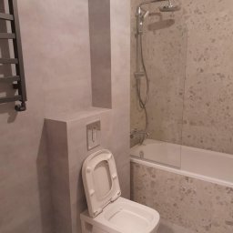 Service4You - Nowoczesna łazienka z wiszącą toaletą z otwartą klapą, kabiną prysznicową za szklaną ścianką i grzejnikiem drabinkowym na ścianie z szarych płytek.