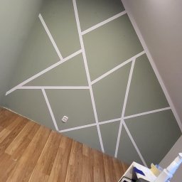 Service4You - Ściana w pokoju na poddaszu, pomalowana na oliwkowy kolor z geometrycznym wzorem z białej taśmy malarskiej, widoczna podłoga z paneli i fragment szafki z narzędziami.