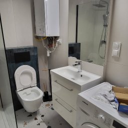 Service4You - Nowoczesna łazienka z białą ceramiką, umywalką z szafką, pralką Samsung i podłogą w stylu lastryko. Na ścianie wisi biały piec gazowy z widocznymi rurami.