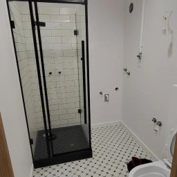 Service4You - Nowoczesna kabina prysznicowa z czarnymi profilami i białymi płytkami w trakcie montażu, widoczne niedokończone instalacje wodne i kanalizacyjne na ścianie, wzorzysta podłoga.