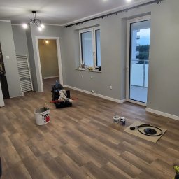 Service4You - Przygotowania do malowania ścian w pokoju w Rzeszowie, widoczne narzędzia malarskie, wiadro z farbą, pędzle i wałki na podłodze z paneli imitujących drewno.