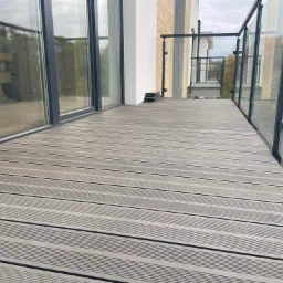 Nowoczesny balkon z szarą deską kompozytową ułożoną poziomo, szklane balustrady z czarnymi ramami, widoczne elementy montażowe na podłodze. W tle budynek z oknami.