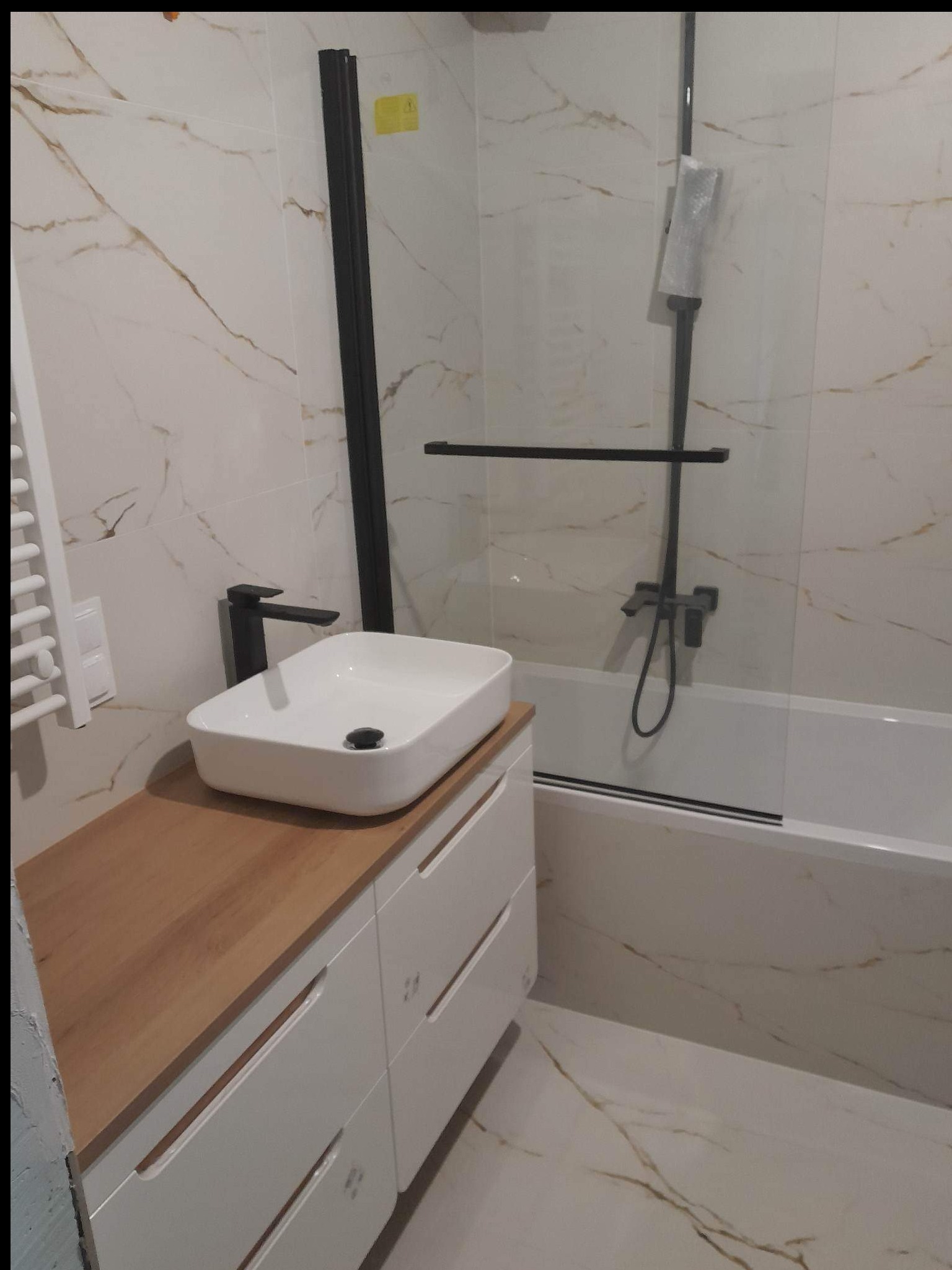 Nowoczesna łazienka z białymi płytkami imitującymi marmur, białą szafką pod umywalką z drewnianym blatem, czarną armaturą i kabiną prysznicową z czarnymi profilami. Na baterii prysznicowej widoczna...