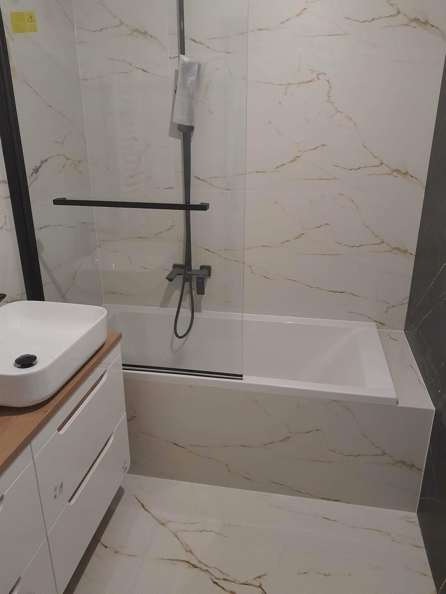 Nowoczesna łazienka z wanną, białymi płytkami imitującymi marmur z wyraźnymi złotymi żyłami, czarną armaturą i szklanym panelem oddzielającym wannę, umywalka nablatowa na białej szafce z drewnianym...