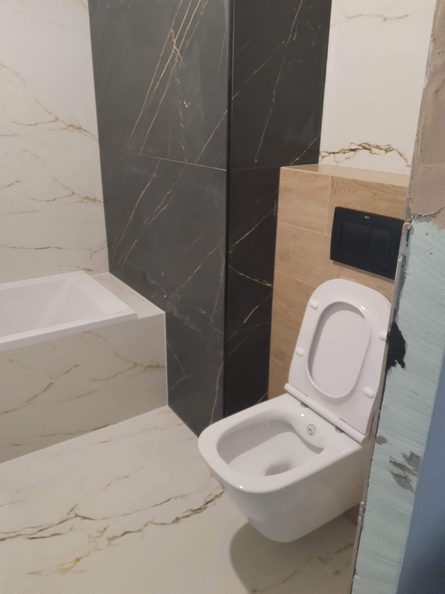 Nowoczesna łazienka z białą wanną, toaletą z deską w górze i płytkami imitującymi marmur w odcieniach bieli i czerni ze złotymi żyłkami, widoczne niedokończone wykończenie ściany.