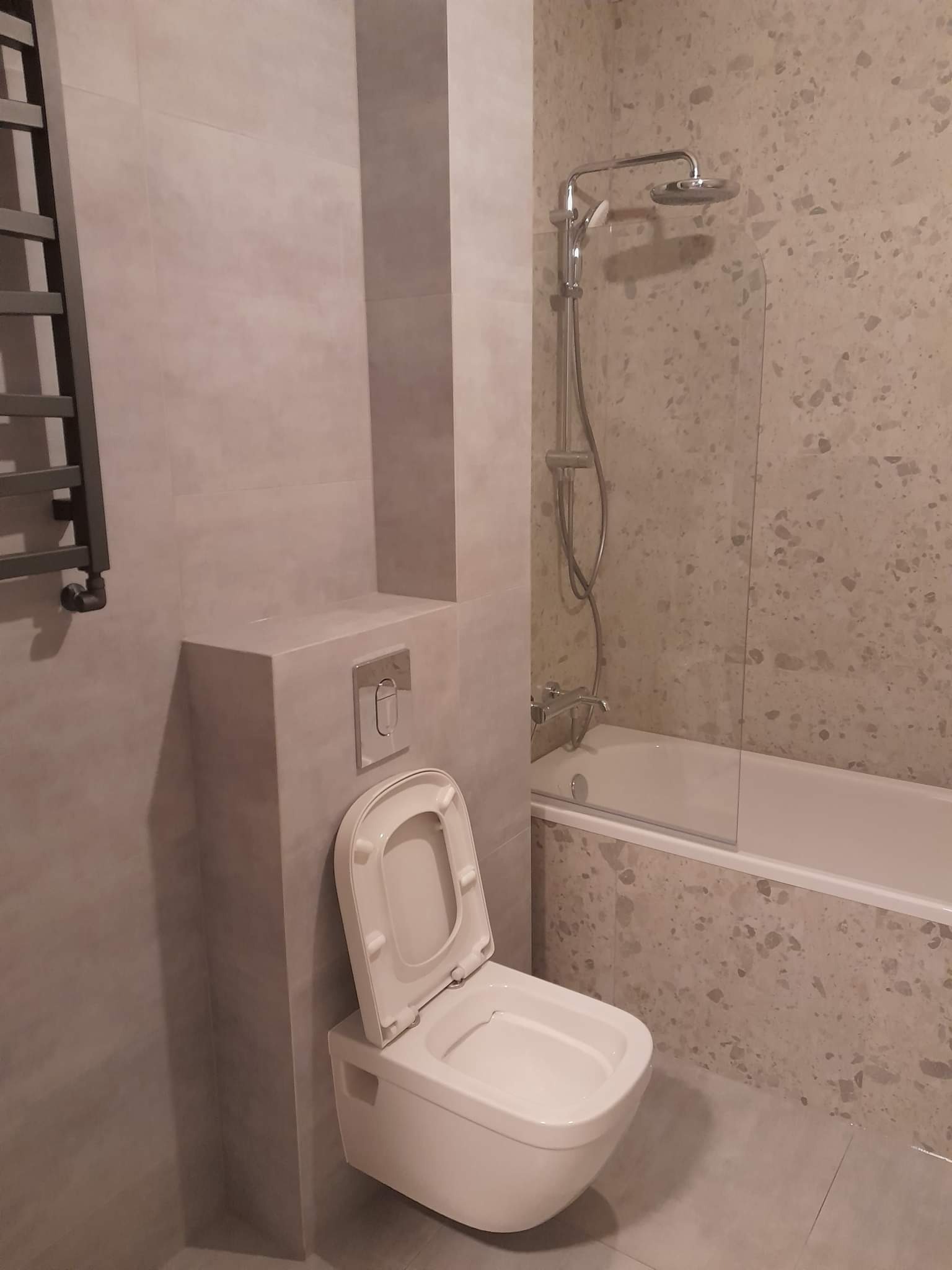 Nowoczesna łazienka z wiszącą toaletą z otwartą klapą, kabiną prysznicową za szklaną ścianką i grzejnikiem drabinkowym na ścianie z szarych płytek.