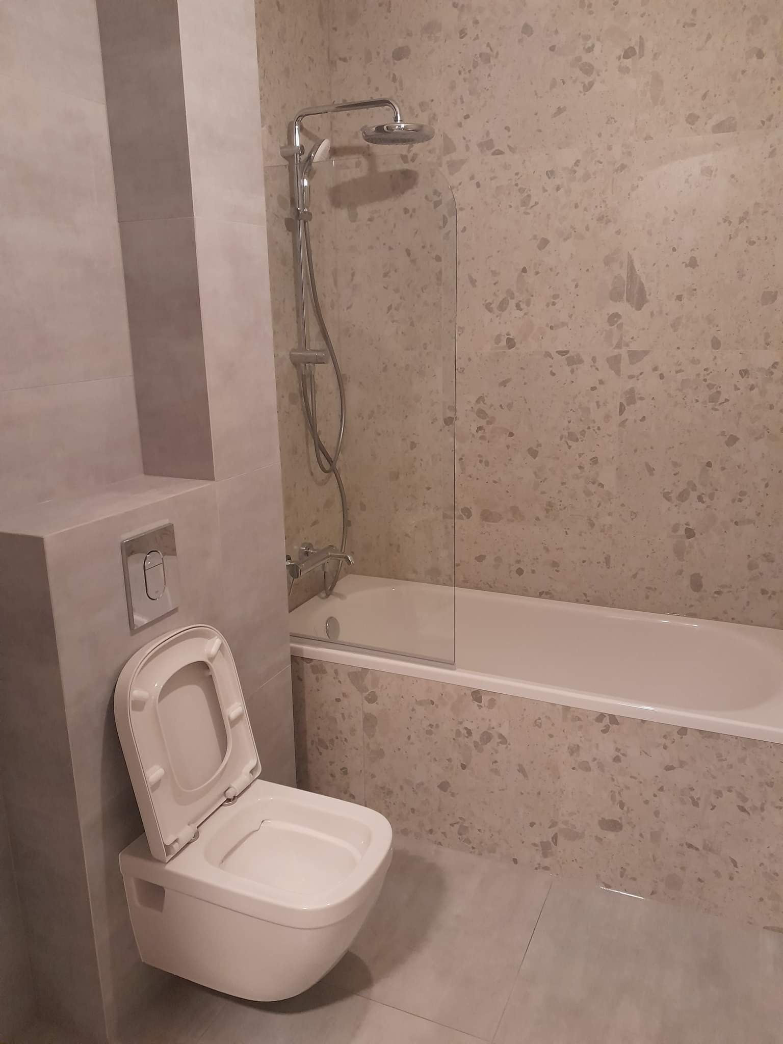 Nowoczesna łazienka z podwieszaną toaletą z otwartą klapą, wanną z parawanem i ścianami wyłożonymi płytkami imitującymi lastryko w odcieniach beżu i szarości.