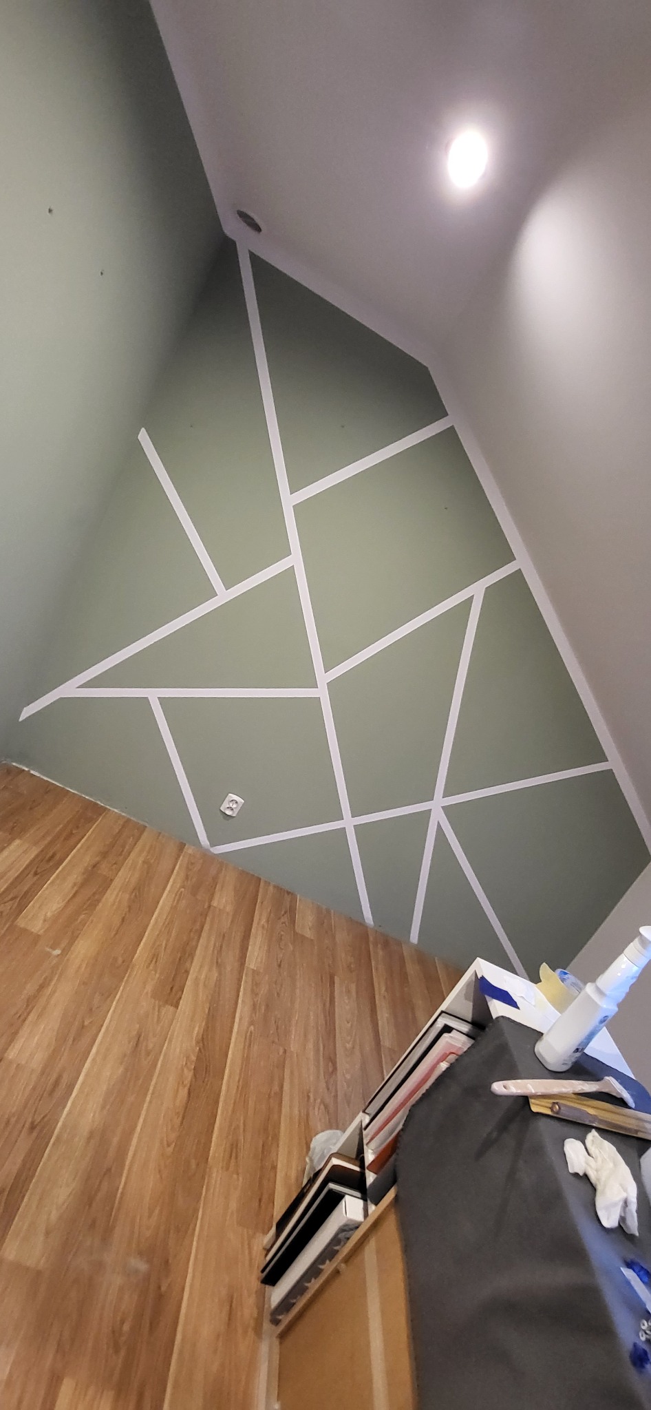 Ściana w pokoju na poddaszu, pomalowana na oliwkowy kolor z geometrycznym wzorem z białej taśmy malarskiej, widoczna podłoga z paneli i fragment szafki z narzędziami.