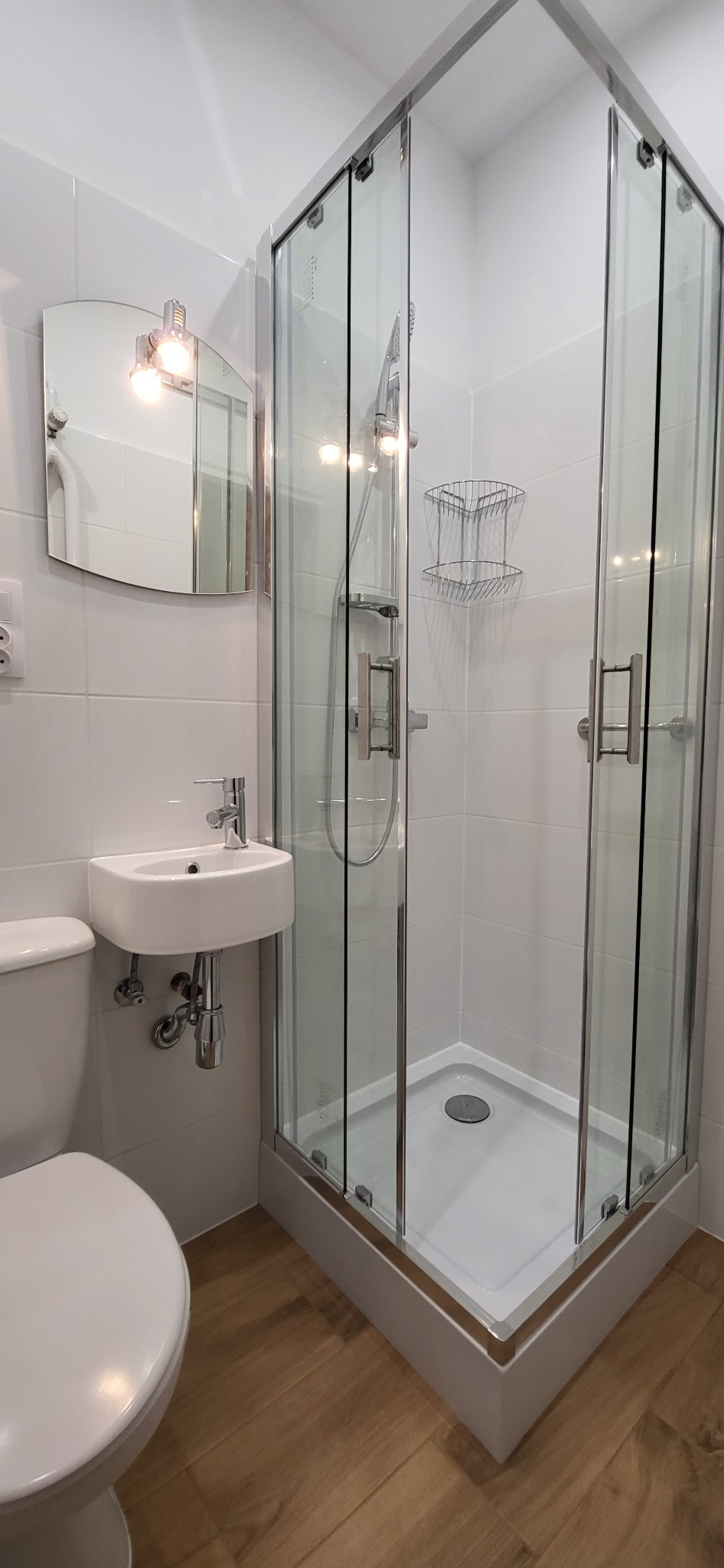 Nowoczesna, mała łazienka z narożną kabiną prysznicową z chromowanymi profilami, podwieszaną umywalką i lustrem z oświetleniem punktowym, wykończona białymi płytkami i drewnopodobną podłogą.
