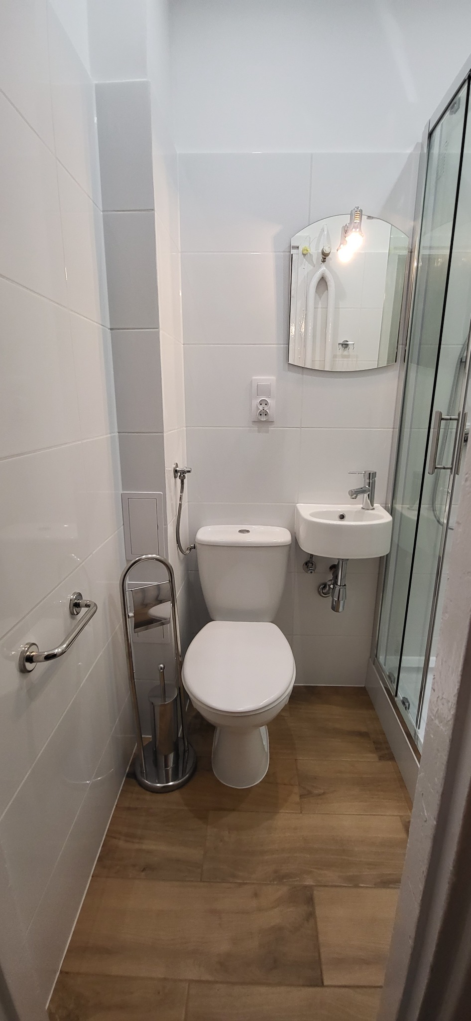 Nowoczesna, mała łazienka z białymi płytkami, drewnopodobną podłogą, wiszącą umywalką i kabiną prysznicową z przezroczystego szkła.