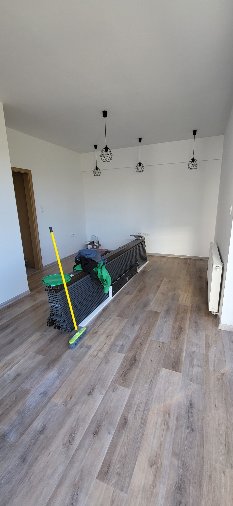 Układanie paneli podłogowych w jasnym pomieszczeniu, widoczna sterta paneli, żółta miotła oparta o panele, oraz trzy czarne, geometryczne lampy wiszące