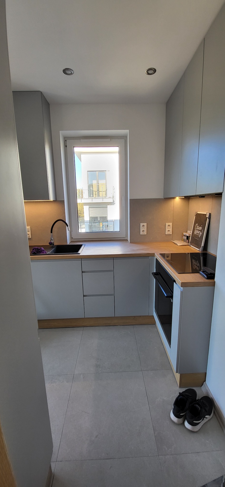 Nowoczesna kuchnia w odcieniach szarości z drewnianym blatem, widok na okno z budynkiem w tle, na podłodze szare płytki i para czarnych butów sportowych.