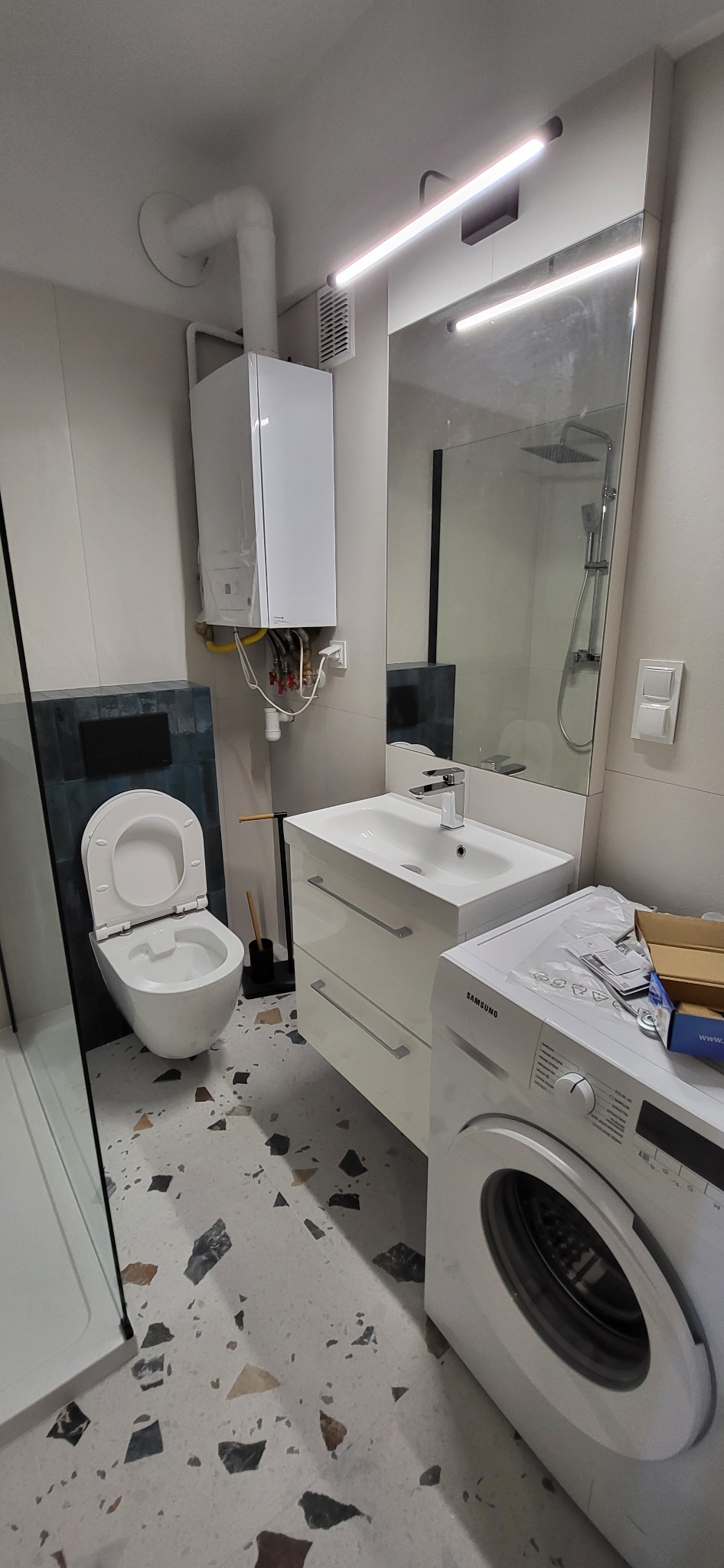 Nowoczesna łazienka z białą ceramiką, umywalką z szafką, pralką Samsung i podłogą w stylu lastryko. Na ścianie wisi biały piec gazowy z widocznymi rurami.