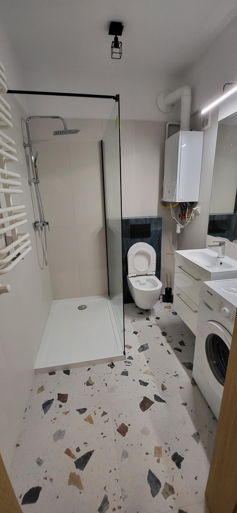 Nowoczesna łazienka z kabiną prysznicową typu walk-in, wiszącą toaletą na tle ciemnej mozaiki, białą umywalką z szafką i pralką Samsung, wykończona płytkami lastryko na podłodze i czarnym...