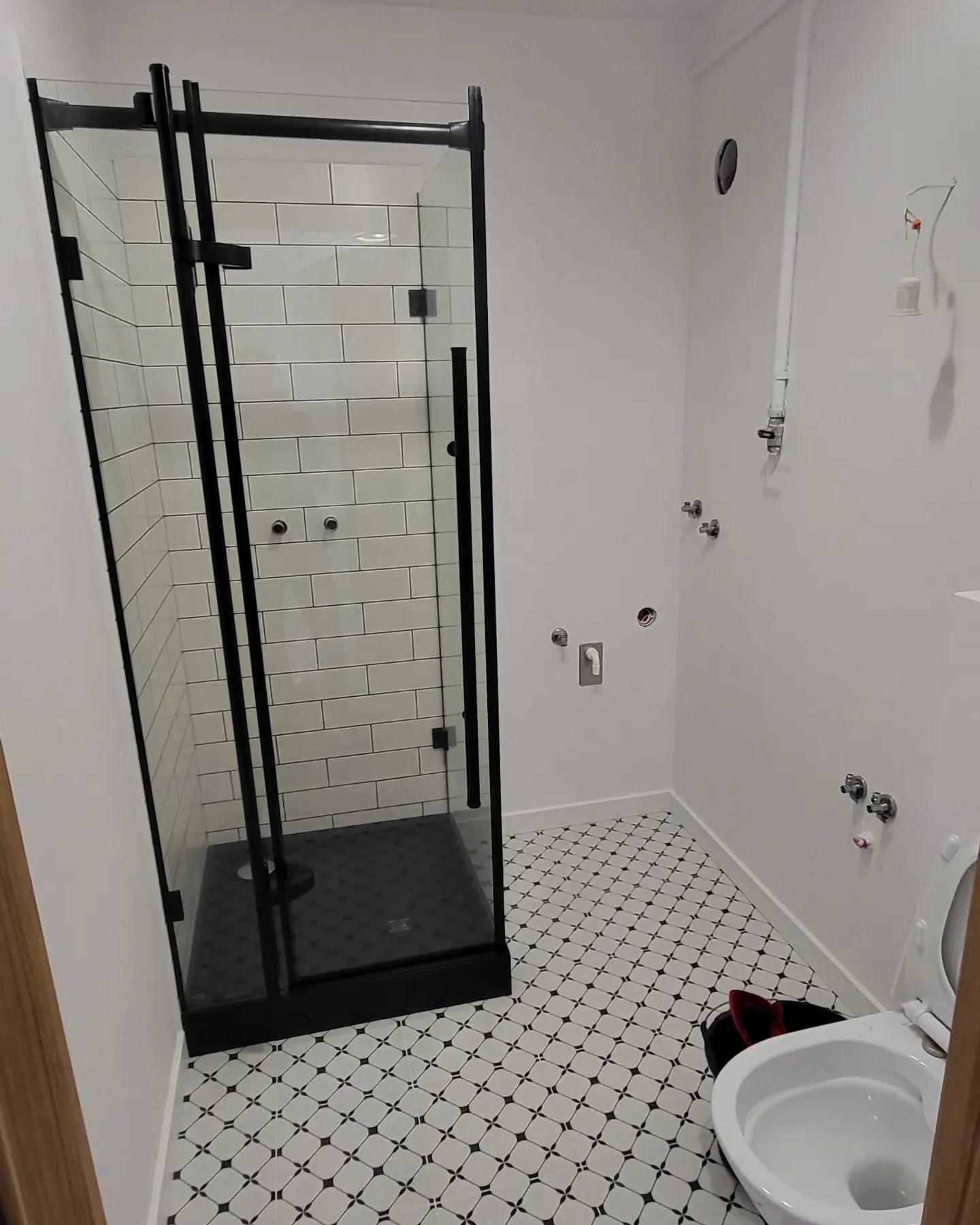 Nowoczesna kabina prysznicowa z czarnymi profilami i białymi płytkami w trakcie montażu, widoczne niedokończone instalacje wodne i kanalizacyjne na ścianie, wzorzysta podłoga.