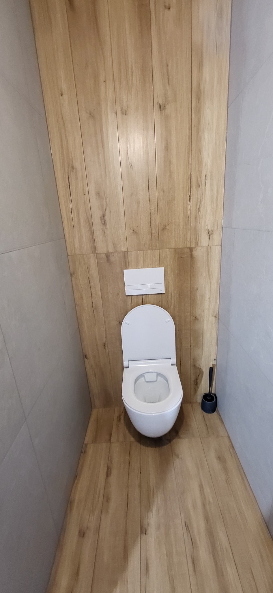 Nowoczesna toaleta wisząca z deską sedesową w kolorze białym, zamontowana na tle ściany wyłożonej płytkami imitującymi drewno, z szarymi płytkami po bokach i czarną szczotką do WC po prawej stronie.