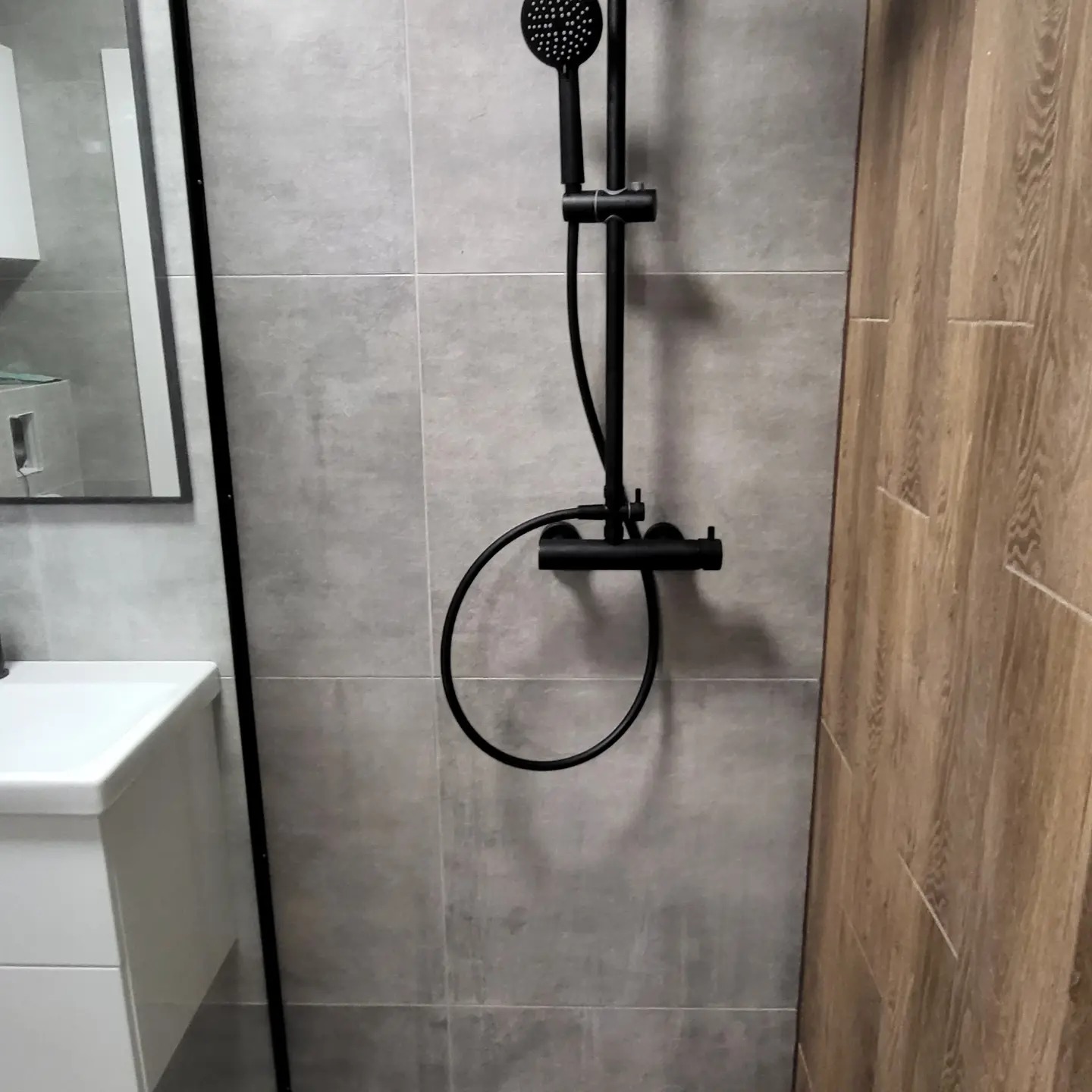 Nowoczesna łazienka z szarymi płytkami, drewnopodobnymi panelami i czarną armaturą prysznicową, widoczna biała umywalka z szafką i lustro w czarnej ramie.