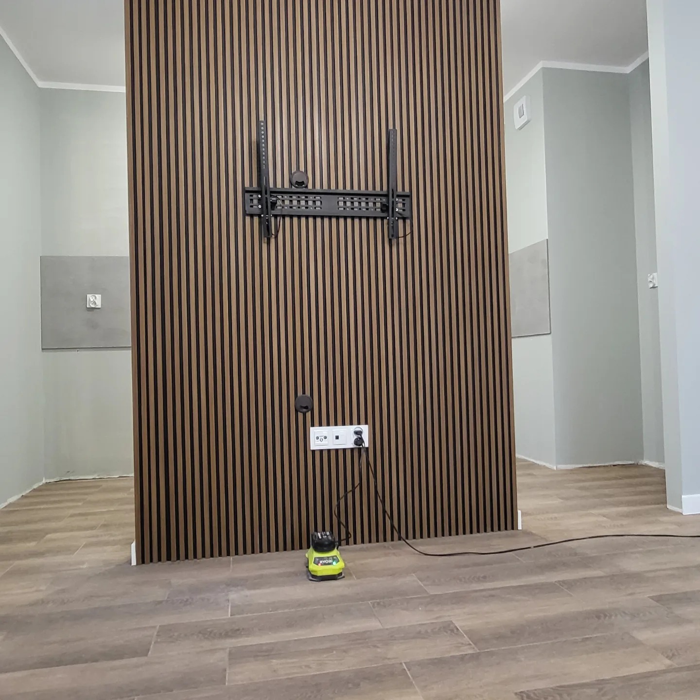Ściana z paneli dekoracyjnych w pionowe pasy w odcieniach brązu i czerni, zamontowany uchwyt do telewizora, gniazdko elektryczne z wpiętą wtyczką, na podłodze leży szlifierka oscylacyjna Ryobi.
