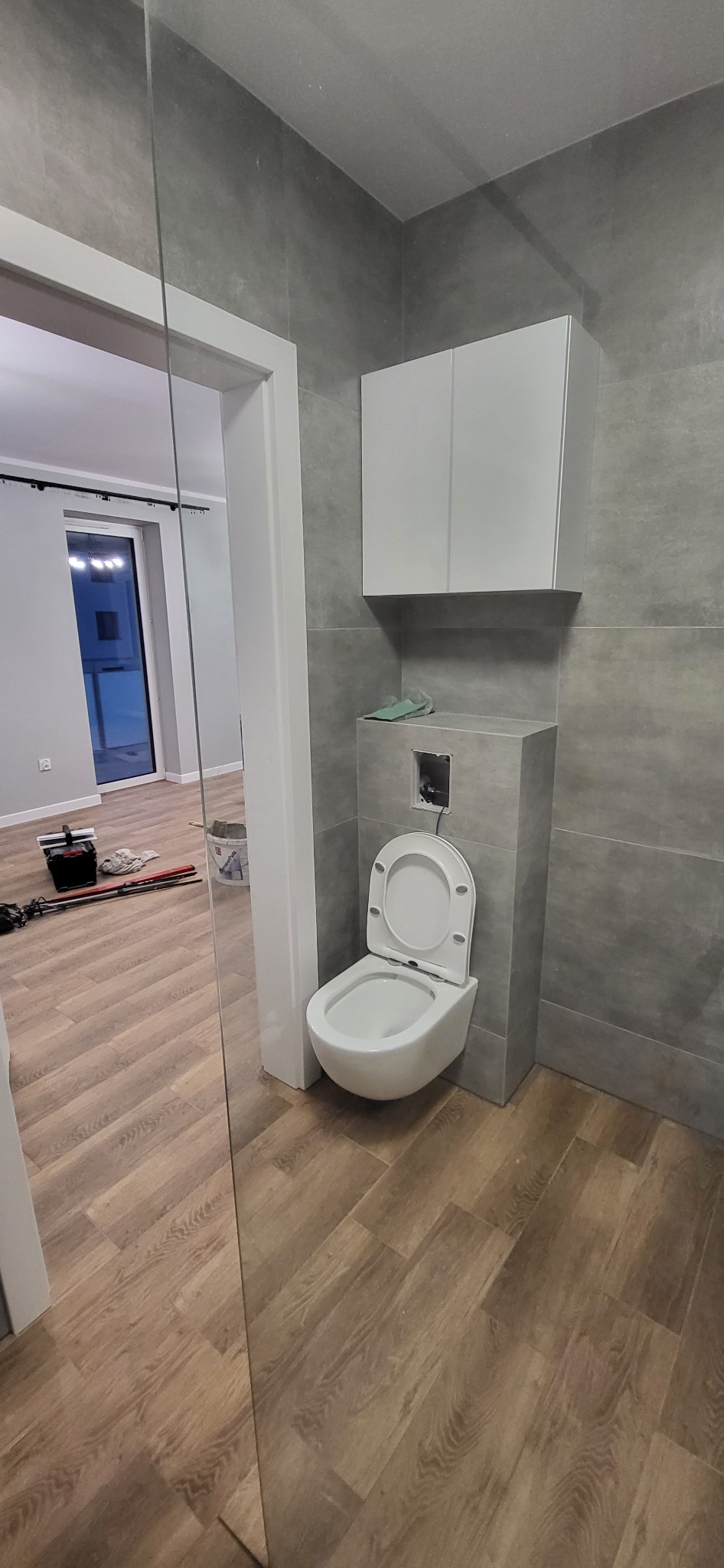 Wisząca toaleta z otwartą klapą, zamontowana na szarej ścianie z płytek, biała szafka wisząca powyżej, widoczne narzędzia i podłoga imitująca drewno w sąsiednim pomieszczeniu, odbicie w lustrze.