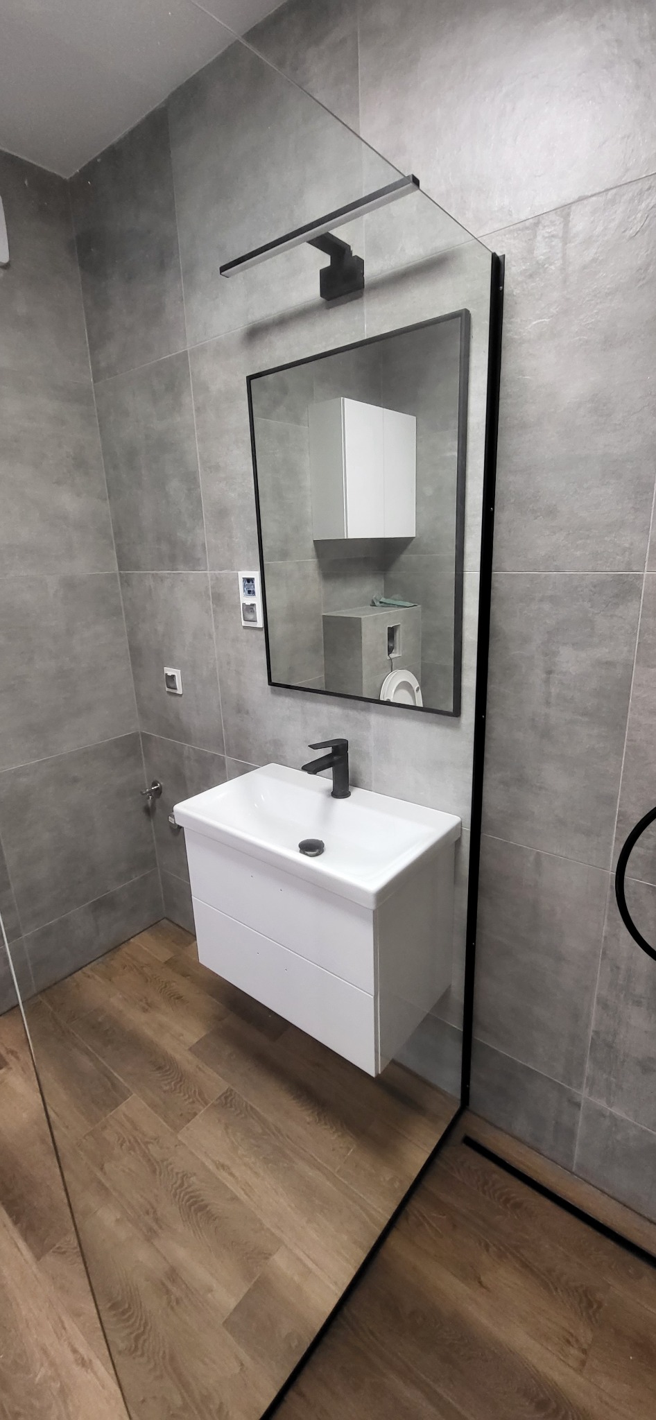 Nowoczesna łazienka z szarą płytką, drewnopodobną podłogą, białą szafką pod umywalką z czarną armaturą i lustrem w czarnej ramie, odbijającym toaletę i szafkę wiszącą.