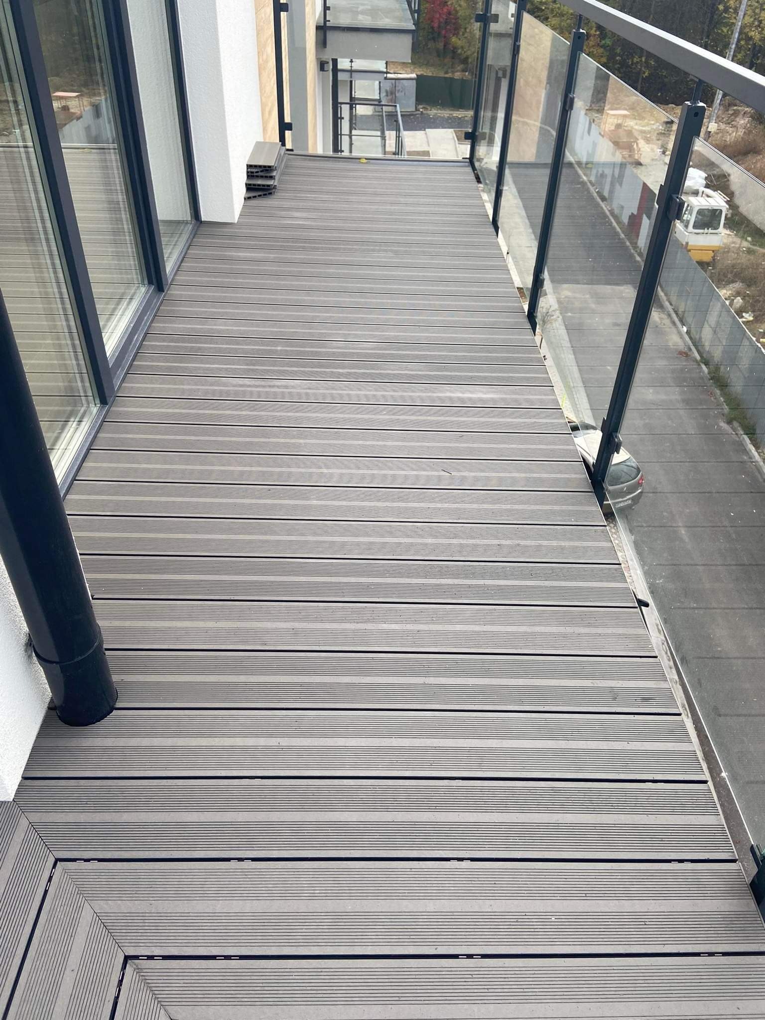 Nowoczesny balkon z szarym, kompozytowym parkietem ułożonym wzdłuż, szklane balustrady z czarnymi ramami, widok na zewnątrz na plac budowy.
