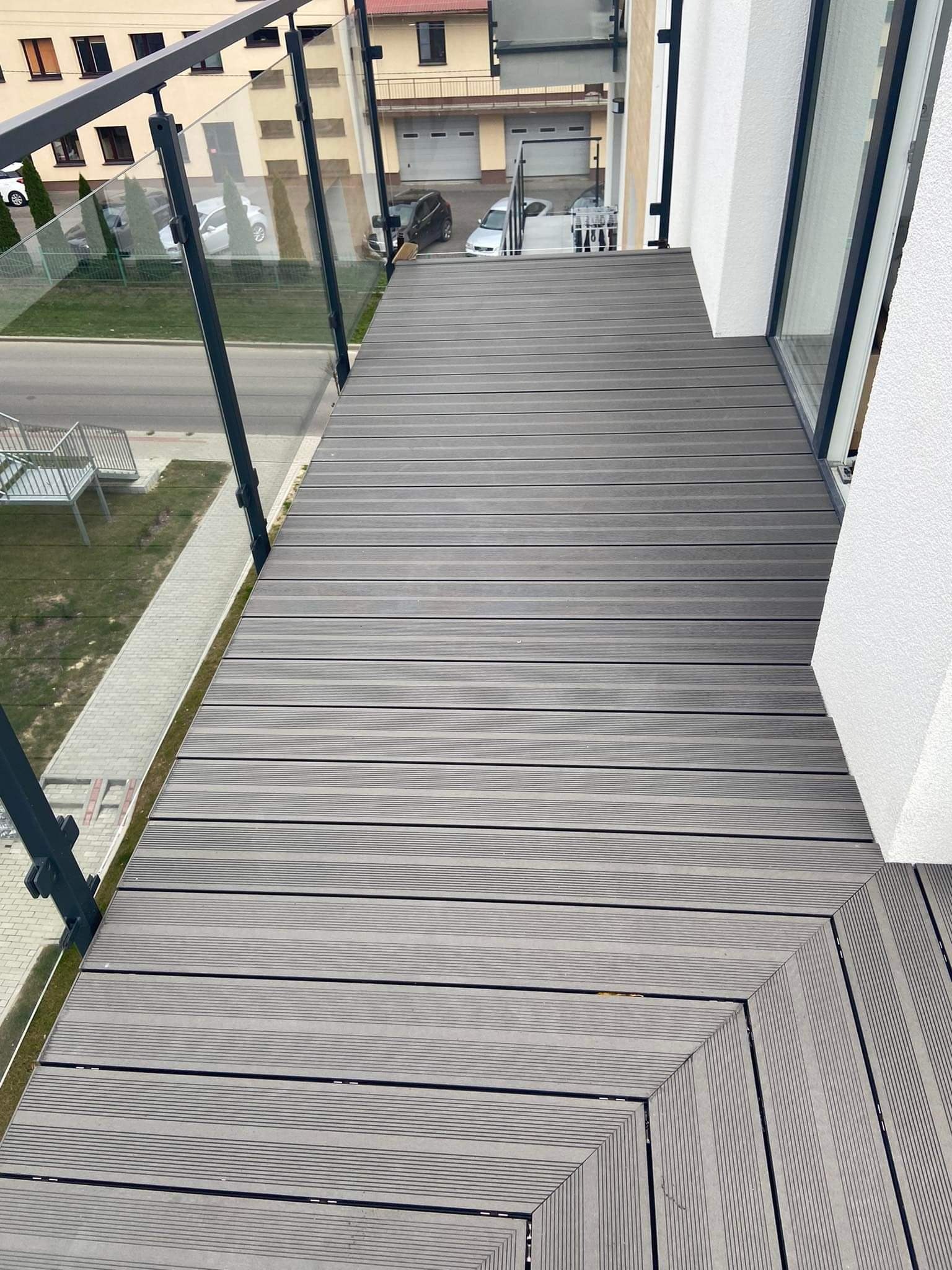 Szare deski kompozytowe ułożone na balkonie z przeszkloną balustradą, widok z góry na fragment podłogi i otoczenie budynku.