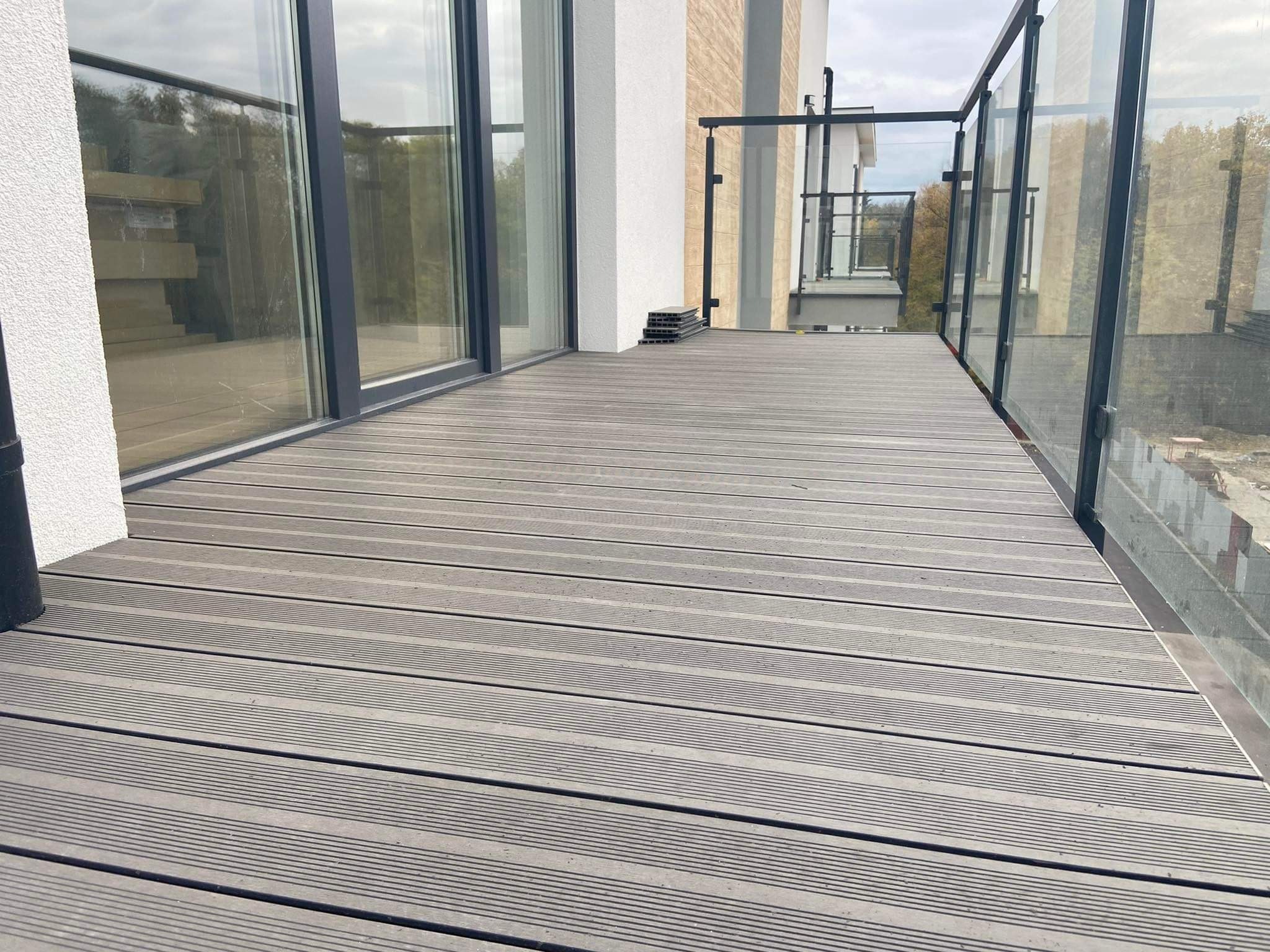 Nowoczesny balkon z szarą deską kompozytową ułożoną poziomo, szklane balustrady z czarnymi ramami, widoczne elementy montażowe na podłodze. W tle budynek z oknami.