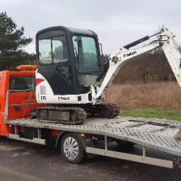 Pomarańczowy samochód transportowy z mini koparką Bobcat E16 na platformie, widok z boku na tle jesiennego krajobrazu.