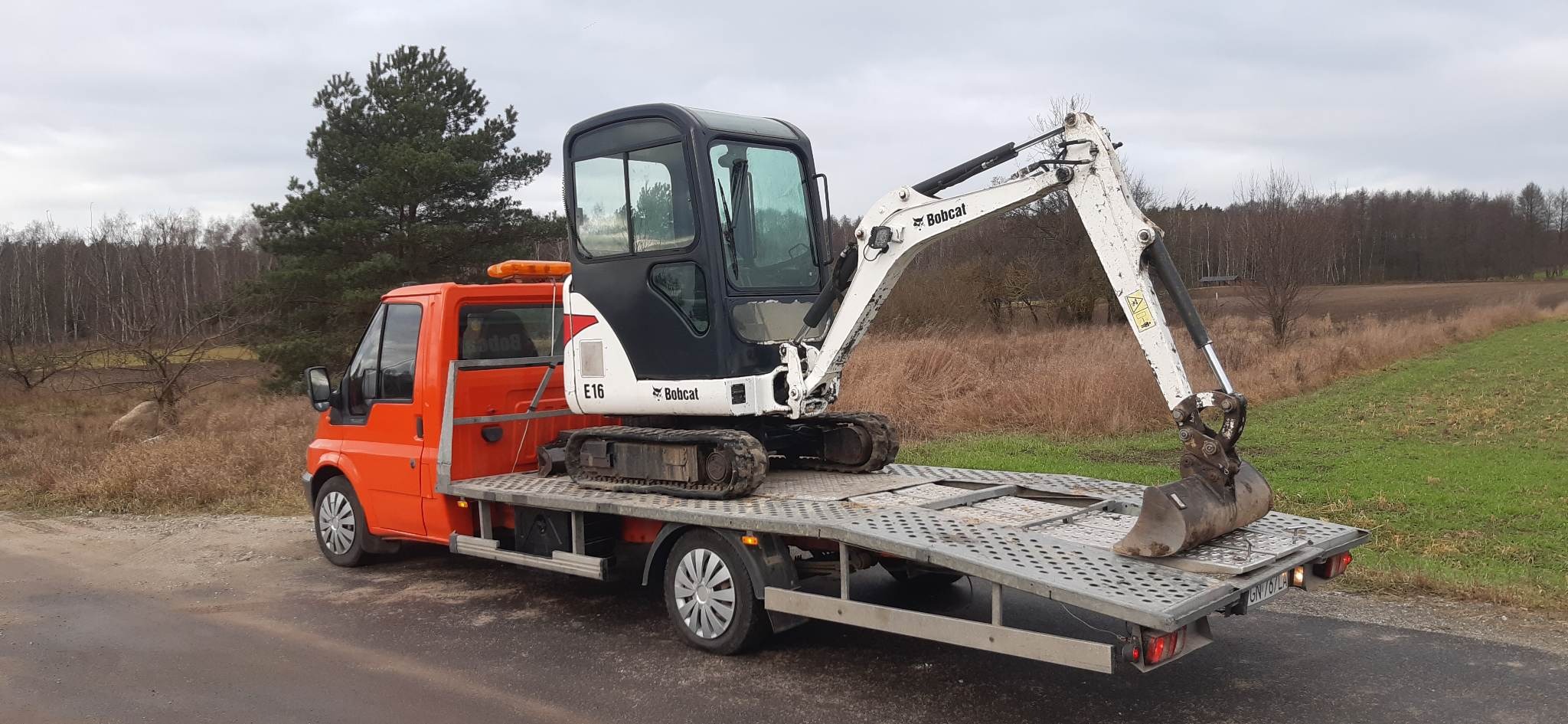 Pomarańczowy samochód transportowy z mini koparką Bobcat E16 na platformie, widok z boku na tle jesiennego krajobrazu.