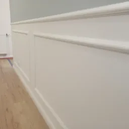 Biała boazeria angielska na ścianie w jasnym wnętrzu, panele podłogowe imitujące drewno, grzejnik na ścianie w tle.