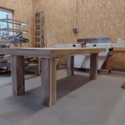 Surowy drewniany stół z grubym blatem i nogami w warsztacie w Narolu, widoczna struktura drewna i maszyny stolarskie w tle.