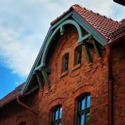 Fragment budynku z czerwonej cegły z widoczną konstrukcją dachu i ozdobnymi elementami drewnianymi w kolorze zielonym, dach pokryty czerwoną dachówką, na tle błękitne niebo.