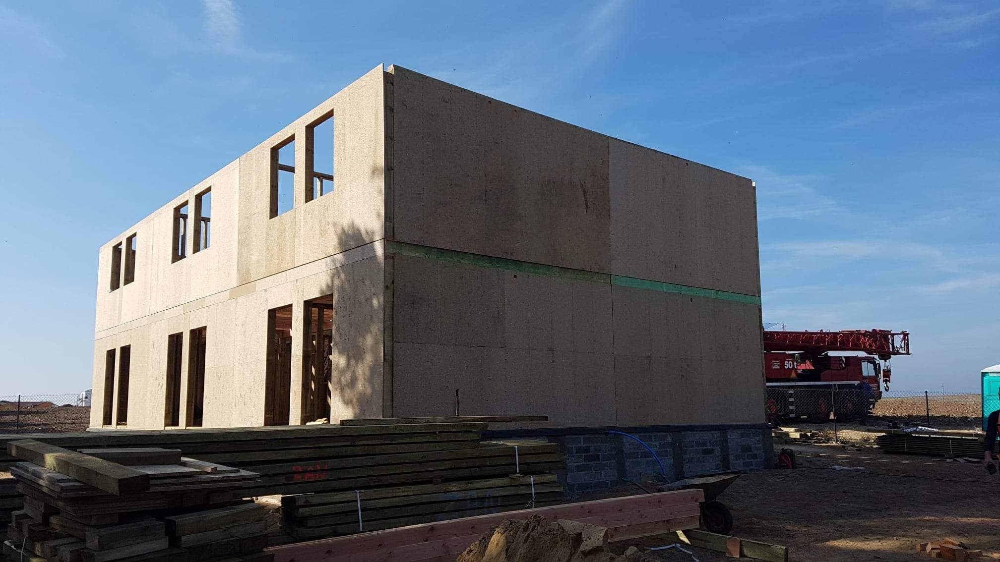 Surowy dwukondygnacyjny budynek w trakcie wznoszenia z drewnianych paneli, widoczny dźwig budowlany w tle i stosy drewna na pierwszym planie.