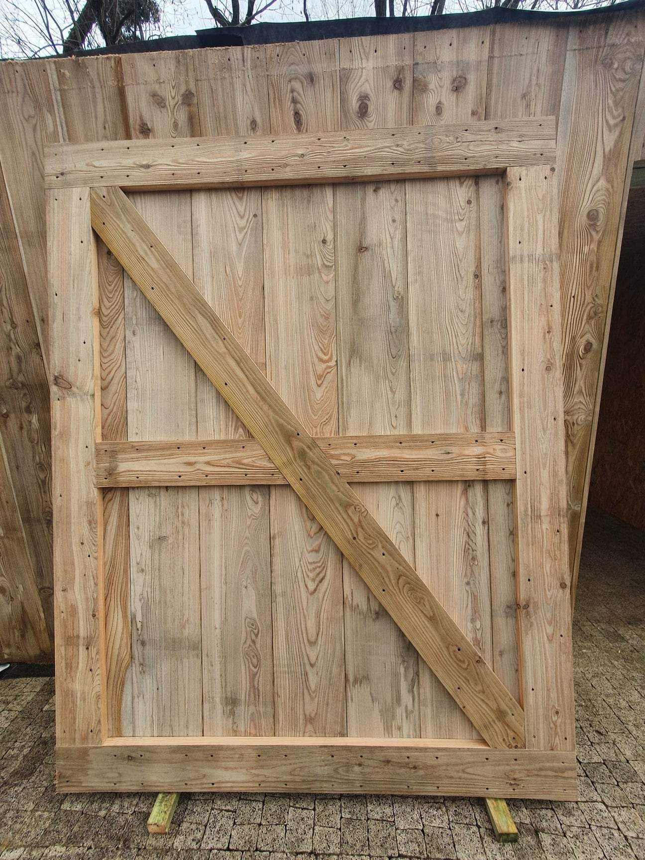 Surowe drewniane elementy konstrukcyjne altany lub domku szkieletowego, z ukośnym wzmocnieniem i widocznymi śladami po wkrętach, leżące na brukowanej powierzchni.