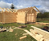 Konstrukcja domu w stanie surowym otwartym, widoczne drewniane ściany i elementy szkieletu dachu, deski na trawie, zielone otoczenie.