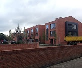 Nowoczesne domy z czerwonej cegły z dużymi oknami i płaskimi dachami w zabudowie szeregowej, widoczne zza niskiego murku z cegły. Na drugim planie drzewa i żółta ciężarówka.