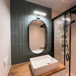 Nowoczesna łazienka: biała umywalka nablatowa na drewnianym blacie, grafitowe płytki na ścianie, czarna rama lustra i kabiny prysznicowej, minimalistyczny design.