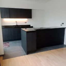 Nowoczesna kuchnia z czarnymi szafkami i wyspą, oświetleniem LED pod szafkami wiszącymi, widoczne fragmenty podłogi z płytek i parkietu.