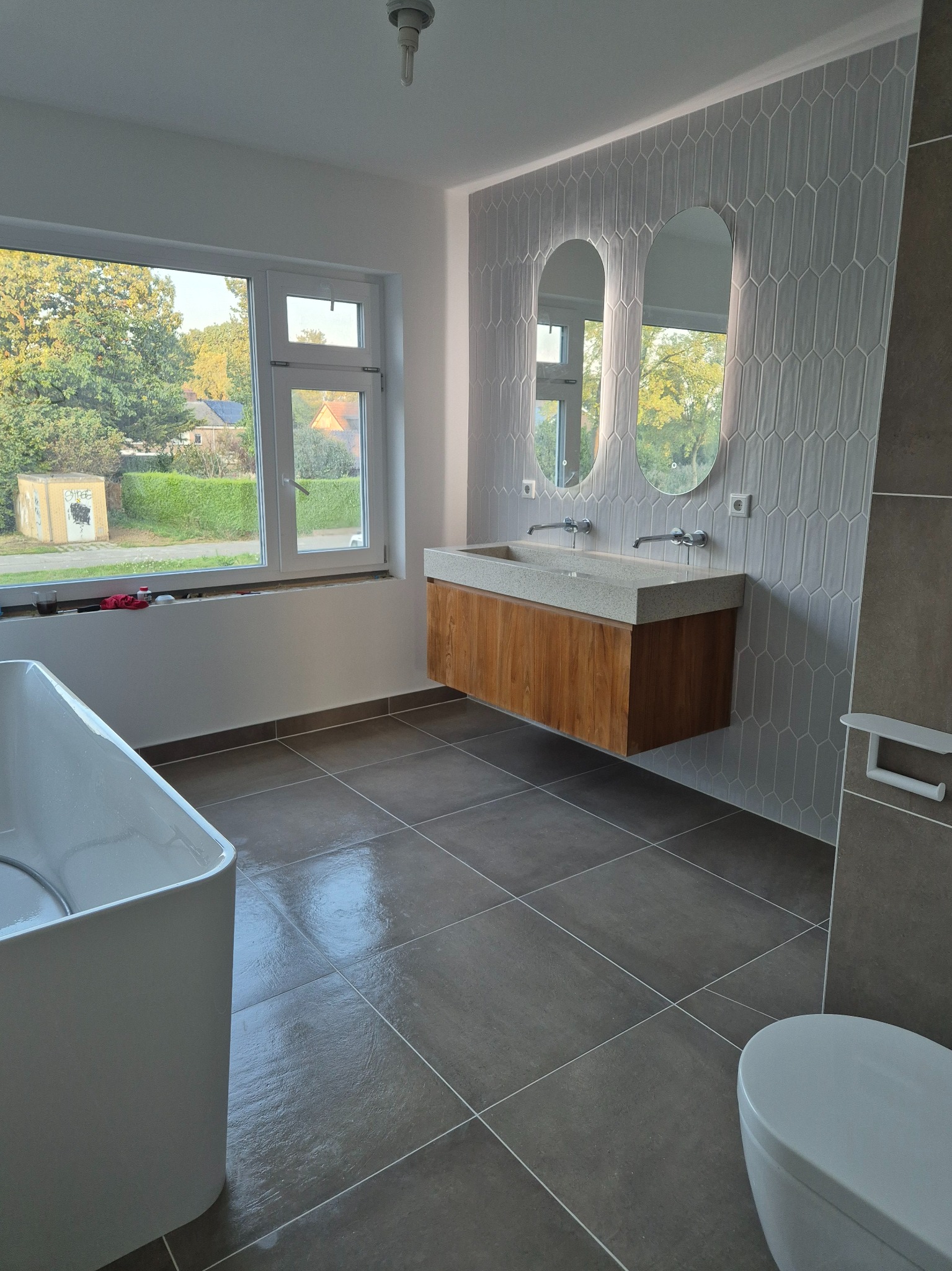 Nowoczesna łazienka z podwójną umywalką na drewnianej szafce, owalnymi lustrami i szarą mozaiką na ścianie, widok na zewnątrz przez okno.