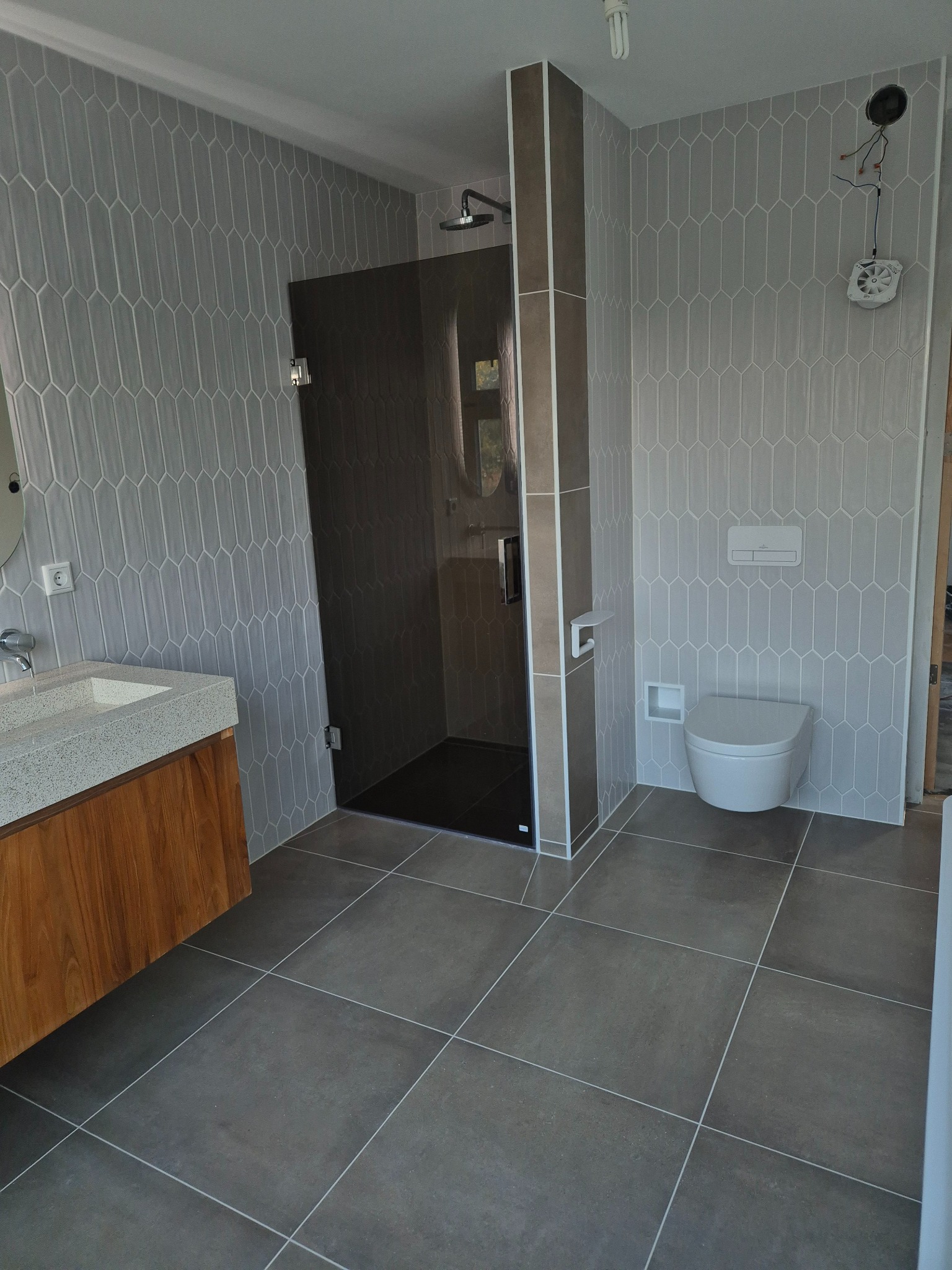 Nowoczesna łazienka po remoncie w Kielcach z kabiną prysznicową typu walk-in, umywalką nablatową z drewnianą szafką, podwieszaną miską WC i szarymi płytkami w kształcie rombów na ścianach.