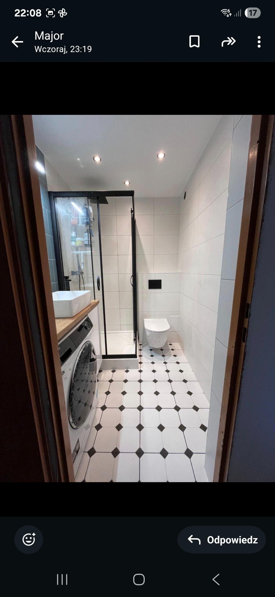 Wyremontowana łazienka z kabiną prysznicową, umywalką na blacie, pralką i biało-czarną podłogą w kratkę. Widok zza framugi drzwi.