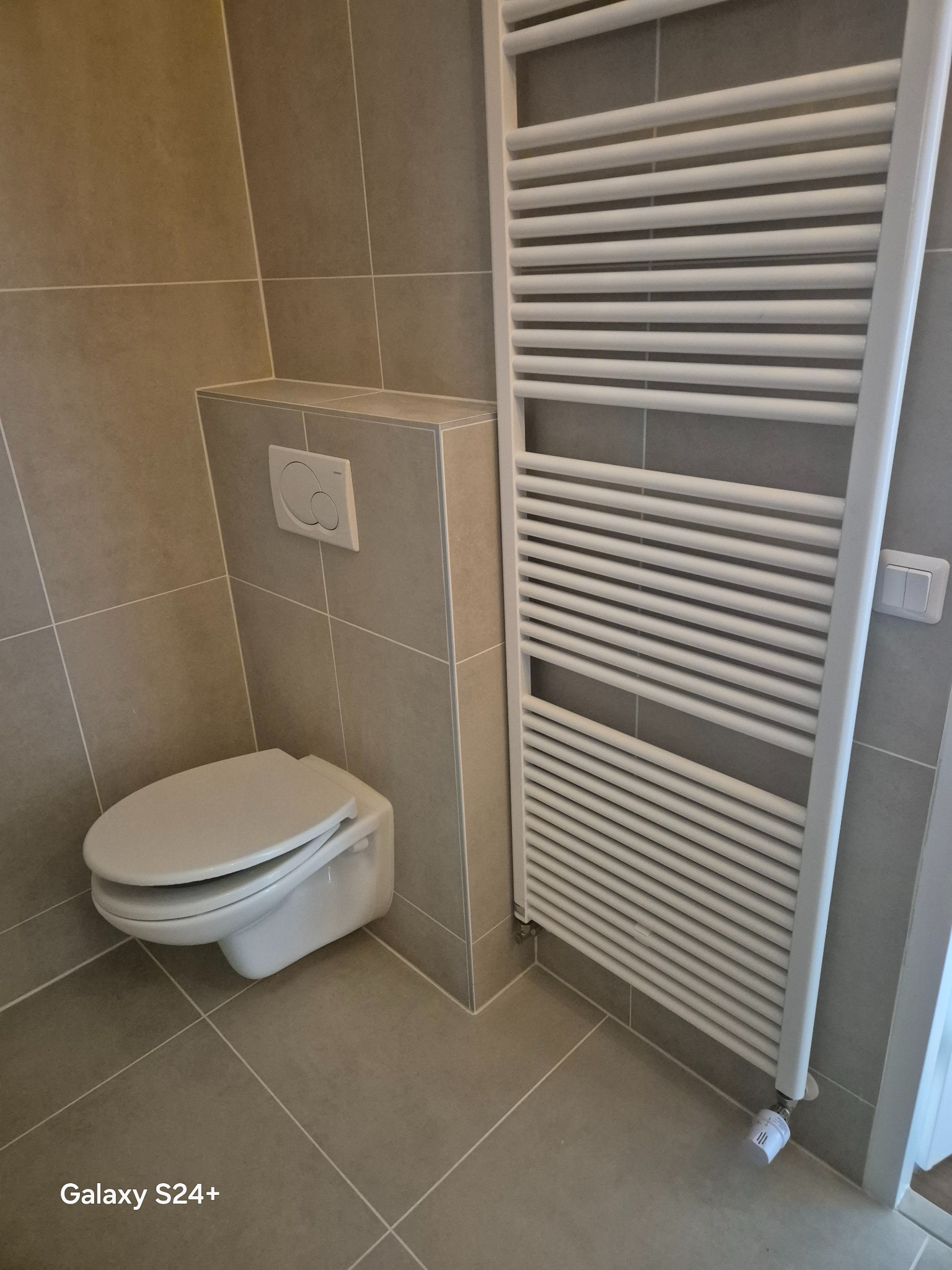 Wykończona łazienka z podwieszaną toaletą i białym grzejnikiem drabinkowym na tle płytek w odcieniach szarości. Widoczny minimalistyczny panel spłuczki.