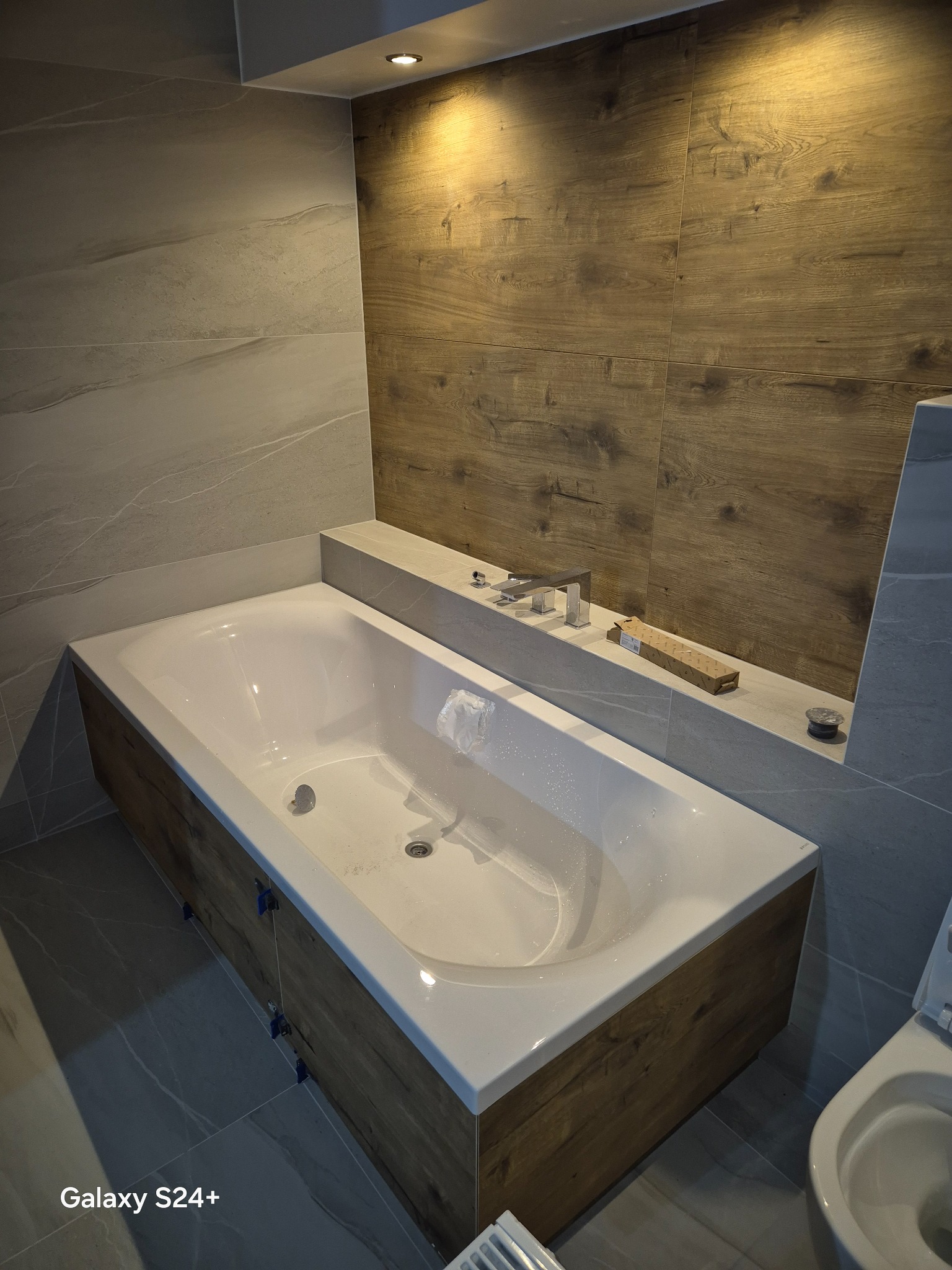 Nowoczesna łazienka po remoncie: wanna obudowana drewnem, szare płytki imitujące marmur na ścianach, drewnopodobne płytki na ścianie, bateria umywalkowa.