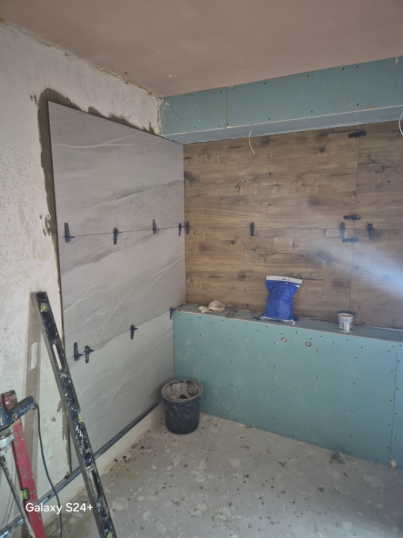 Układanie płytek imitujących marmur na ścianie łazienki, z użyciem poziomicy i klipsów. Widoczne płyty gipsowe i narzędzia glazurnicze. Prace w toku.