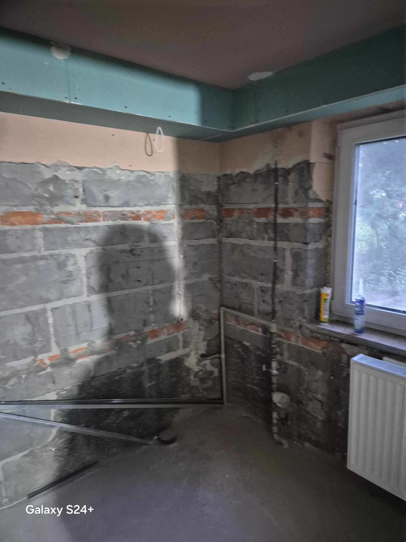 Surowa ściana z cegły i betonu w trakcie remontu łazienki, widoczne rury i grzejnik, przygotowanie do dalszych prac wykończeniowych.