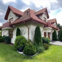 Dwupiętrowy dom z czerwonym dachem, balkonem i zadbanym ogrodem z krzewami i trawnikiem, widziany z perspektywy ogrodu.