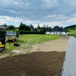 Sun Grass - Przygotowanie terenu pod trawnik: wyrównana, ciemna ziemia, piasek, zielony trawnik, mini koparka i dom w tle.